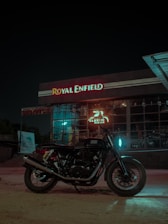 A motorcycle is parked in front of a Royal Enfield dealership at night. The building features large glass windows and illuminated signage displaying the name 'Royal Enfield' in red and white. The area is dimly lit with a glowing blue light reflecting on the motorcycle.