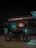 A motorcycle is parked in front of a Royal Enfield dealership at night. The building features large glass windows and illuminated signage displaying the name 'Royal Enfield' in red and white. The area is dimly lit with a glowing blue light reflecting on the motorcycle.