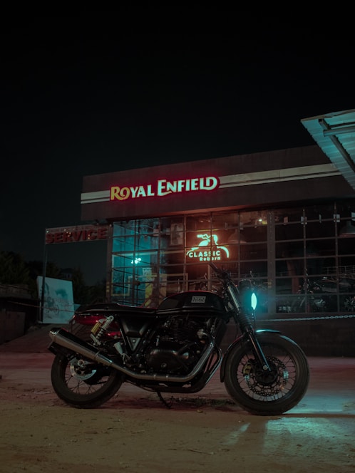 A motorcycle is parked in front of a Royal Enfield dealership at night. The building features large glass windows and illuminated signage displaying the name 'Royal Enfield' in red and white. The area is dimly lit with a glowing blue light reflecting on the motorcycle.