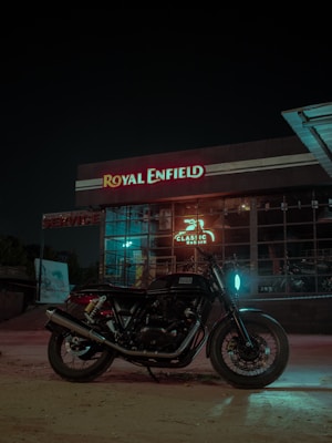 A motorcycle is parked in front of a Royal Enfield dealership at night. The building features large glass windows and illuminated signage displaying the name 'Royal Enfield' in red and white. The area is dimly lit with a glowing blue light reflecting on the motorcycle.