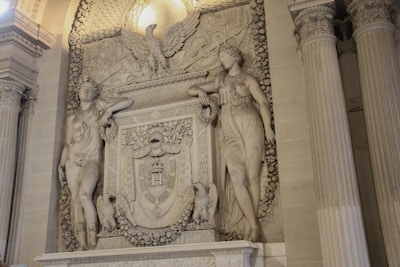 A detailed stone relief featuring two classical figures standing on either side of a central heraldic shield. The shield is adorned with intricate carvings, including laurel wreaths and an eagle perched at the top. The background is embellished with ornate floral decorations. Two imposing Corinthian columns flank the scene.