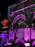 An exterior view of a historical building with a prominent arched window, illuminated in purple and pink lights. The sign for Hard Rock Cafe Bengaluru is brightly lit in red, attached to the stone façade with intricate architectural details.