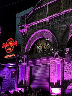 An exterior view of a historical building with a prominent arched window, illuminated in purple and pink lights. The sign for Hard Rock Cafe Bengaluru is brightly lit in red, attached to the stone façade with intricate architectural details.
