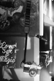 A black and white photograph features a vintage film camera hanging from a strap on a wooden surface. To the left, a sign with stylized text reads 'Love Sex & Magic'. Behind the camera and sign, part of a painting or art piece is partially visible, depicting abstract objects and shapes.