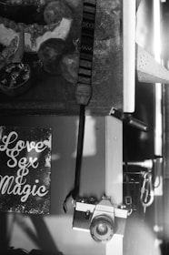 A black and white photograph features a vintage film camera hanging from a strap on a wooden surface. To the left, a sign with stylized text reads 'Love Sex & Magic'. Behind the camera and sign, part of a painting or art piece is partially visible, depicting abstract objects and shapes.