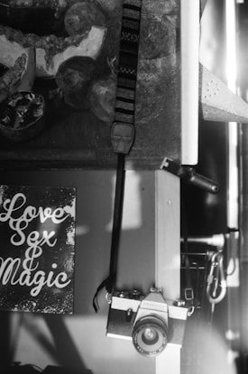 A black and white photograph features a vintage film camera hanging from a strap on a wooden surface. To the left, a sign with stylized text reads 'Love Sex & Magic'. Behind the camera and sign, part of a painting or art piece is partially visible, depicting abstract objects and shapes.