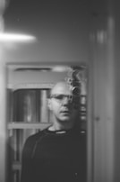 A black and white photograph of a person looking into a mirror, capturing their reflection. The reflection prominently features a vintage camera positioned in front of the person's face. The setting appears to be indoors with soft lighting.