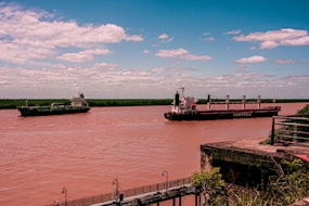 Two large cargo ships navigate a wide river under a clear, lightly clouded sky. The riverbank features some greenery and a rusted metal railing in the foreground. The larger ship, labeled 'CAMPBELL,' is moving along with the other vessel alongside.