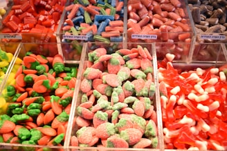 An assortment of colorful gummy candies displayed in clear containers, including red licorice pieces, blue and green twisted sticks, red gummies with white tips, and red and green sugar-coated strawberries. Each container holds a different type of candy, showcasing a variety of shapes and colors.