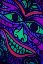 A vibrant, abstract drawing depicting a face with exaggerated eyes and a wide grin, featuring swirling patterns. The artwork is rendered in vivid neon colors including green, purple, blue, and pink against a dark background. The style is bold and expressive, using heavy black outlines to define facial features and intricate designs.