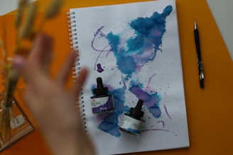 A spiral-bound notebook is open to a page filled with abstract watercolor patterns featuring blue and purple hues. Two small bottles of Ecoline liquid watercolor are resting on the notebook. A hand is reaching toward the left side of the frame, and a black pen is positioned next to the notebook. The surface beneath is a vibrant orange color.