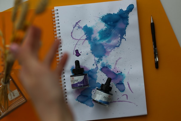 A spiral-bound notebook is open to a page filled with abstract watercolor patterns featuring blue and purple hues. Two small bottles of Ecoline liquid watercolor are resting on the notebook. A hand is reaching toward the left side of the frame, and a black pen is positioned next to the notebook. The surface beneath is a vibrant orange color.