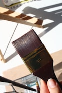 A close-up view of a person holding a large paintbrush with the number 50 engraved on it. The background features a wooden ladder casting shadows on a white surface.