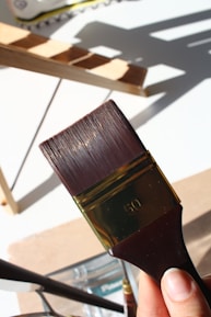 A close-up view of a person holding a large paintbrush with the number 50 engraved on it. The background features a wooden ladder casting shadows on a white surface.