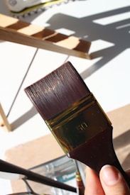 A close-up view of a person holding a large paintbrush with the number 50 engraved on it. The background features a wooden ladder casting shadows on a white surface.