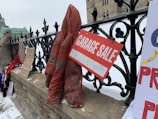 A collection of protest signs and a red coat are displayed on a stone fence outside a large building with Gothic architecture. One sign reads 'GARAGE SALE' with 'TRUDEAU'S HOUSE' written below. The scene is set in snowy weather, and there are multiple handwritten signs placed along the fence.