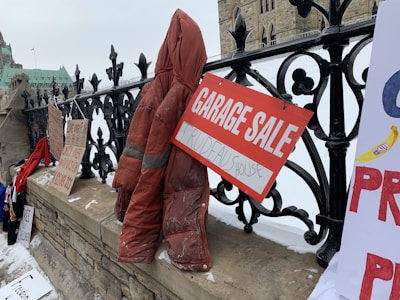 A collection of protest signs and a red coat are displayed on a stone fence outside a large building with Gothic architecture. One sign reads 'GARAGE SALE' with 'TRUDEAU'S HOUSE' written below. The scene is set in snowy weather, and there are multiple handwritten signs placed along the fence.