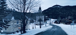 A serene winter village scene with snow covering the landscape. A road curves gently through the village, lined with snow-dusted houses. A church with a steeple stands prominently, framed by bare trees. In the background, forested hills and wind turbines are visible under a muted, overcast sky.