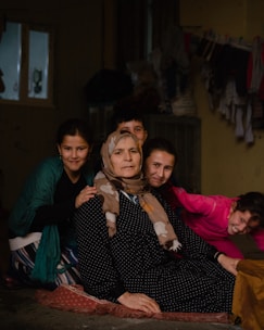 An elderly woman wearing a patterned headscarf and polka dot dress sits on a mat with four smiling children around her. The setting appears to be indoors, possibly a modest home with clothes hanging to dry in the background.