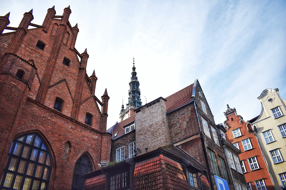 The image depicts a cityscape with three distinct buildings, each showcasing unique architectural styles and colors. On the left side is a red brick building with a pointed roof, featuring multiple arched windows that add to its charm. In the center stands another red brick building with a flat roof, also adorned with numerous arched windows.

On the right side of the image, there's a white building with a pointed roof and a tower on top. The tower is part of a larger structure, which has a flat roof and multiple arched windows as well.