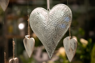A large, textured metallic heart hangs centrally, surrounded by smaller wooden hearts suspended by twine. The background is softly blurred with hints of greenery and warm lighting, creating a serene atmosphere.