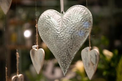 A large, textured metallic heart hangs centrally, surrounded by smaller wooden hearts suspended by twine. The background is softly blurred with hints of greenery and warm lighting, creating a serene atmosphere.