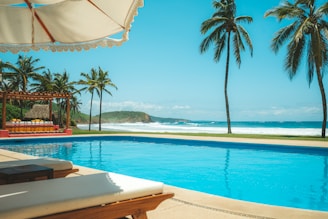 A scenic beachside area with a clear blue swimming pool in the foreground. Tall palm trees and a shaded seating area with sunbeds create a tropical ambiance. The ocean waves gently crash against the shore in the background under a clear, sunny sky.