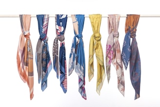 A row of seven colorful and patterned scarves neatly tied onto a horizontal pole. Each scarf features distinct designs, including floral, abstract, and geometric patterns with a variety of colors such as blue, pink, yellow, and brown.