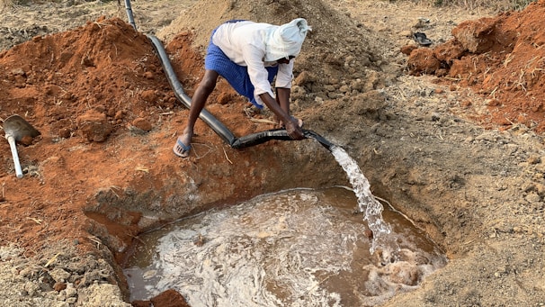A person is using a hose to fill a shallow pit with water in a rural, earthy setting. The ground is composed of reddish-brown soil, and the person is dressed in a white headwrap and blue shorts, standing near the water source.