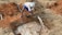 A person is using a hose to fill a shallow pit with water in a rural, earthy setting. The ground is composed of reddish-brown soil, and the person is dressed in a white headwrap and blue shorts, standing near the water source.