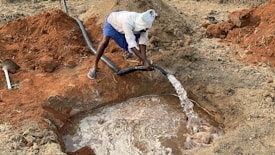 A person is using a hose to fill a shallow pit with water in a rural, earthy setting. The ground is composed of reddish-brown soil, and the person is dressed in a white headwrap and blue shorts, standing near the water source.