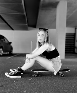 A black and white image of a young person with long hair sitting on a skateboard, dressed in a sleeveless top, denim shorts, and high-top sneakers. The setting appears to be in a parking garage with a parked car visible in the background.