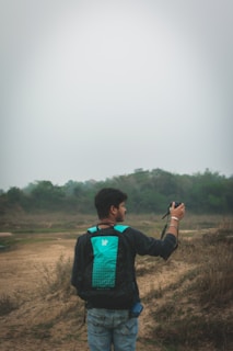 A person with a backpack is taking a photo in a natural setting. The landscape is open with sparse vegetation and a cloudy sky. The person is wearing casual clothing and appears to be focused on the camera held in one hand.