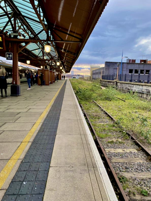 A train station platform features a modern, glass-paneled roof and a long stretch of concrete beside railway tracks. Several people stand on the platform, with a train parked on the left. The sky is partly cloudy with a hint of sunset in the background.