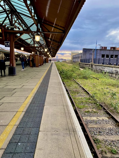 A train station platform features a modern, glass-paneled roof and a long stretch of concrete beside railway tracks. Several people stand on the platform, with a train parked on the left. The sky is partly cloudy with a hint of sunset in the background.