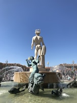 A large sculpture of a male figure is featured, set against a clear blue sky. Water fountains spray in various directions around the statue, which is surrounded by smaller sculptures of aquatic creatures and human figures. Buildings with red roofs can be seen in the background.