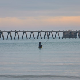 A person stands waist-deep in calm ocean waters holding a fishing rod. In the background, an old wooden pier stretches across the horizon under a softly colored sky.