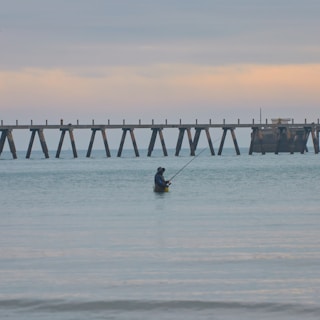 A person stands waist-deep in calm ocean waters holding a fishing rod. In the background, an old wooden pier stretches across the horizon under a softly colored sky.