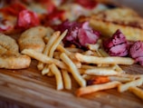 A close-up of a variety of foods including crispy golden French fries, pieces of roasted meat, and baked goods like chicken tenders and pizza slices displayed on a wooden cutting board.