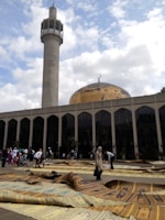 A large mosque with a golden dome and a tall minaret. People gather outside the building on decorative prayer mats spread across the ground. The architecture features tall, arched windows, and the sky is partly cloudy.