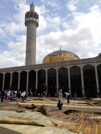 A large mosque with a golden dome and a tall minaret. People gather outside the building on decorative prayer mats spread across the ground. The architecture features tall, arched windows, and the sky is partly cloudy.