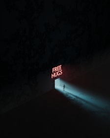 A person stands in a doorway with neon red letters spelling out 'FREE HUGS' above. The surrounding area is dark, with a dim blue light emanating from the door, casting a shadow of the figure onto the floor.