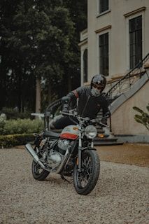A person wearing a helmet and protective gear is sitting on a Royal Enfield motorcycle parked in a gravel area. The background includes a large building with multiple windows and a staircase, surrounded by greenery.