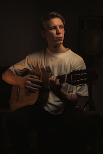 A person is seated playing a classical guitar, with soft, dramatic lighting focusing on him. The background is dark, creating a moody and intimate atmosphere. He is wearing a plain white shirt and has a contemplative expression.