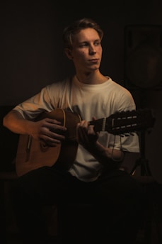 A person is seated playing a classical guitar, with soft, dramatic lighting focusing on him. The background is dark, creating a moody and intimate atmosphere. He is wearing a plain white shirt and has a contemplative expression.