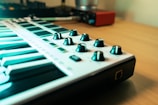A close-up view of a MIDI keyboard with several black knobs on a light-colored wooden desk. The background features an audio interface and a blurred environment, suggesting a music production setup.