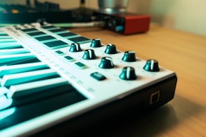 A close-up view of a MIDI keyboard with several black knobs on a light-colored wooden desk. The background features an audio interface and a blurred environment, suggesting a music production setup.