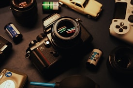 A flat lay photograph features a vintage Nikon camera surrounded by several film rolls, an old toy car, a game controller, and additional camera lenses. The setup is on a dark surface with a warm, nostalgic lighting.
