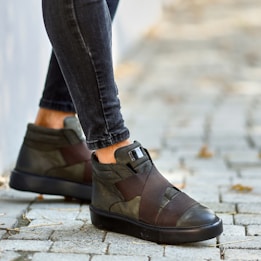 A pair of stylish green high-top sneakers with black soles and broad brown straps is worn by a person wearing skinny dark jeans. The person is walking on a paved pathway with some scattered dry leaves.