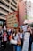 A group of protesters on a city street hold signs with messages about climate change. The central figure carries a megaphone and a sign that reads 'The greatest threat to our planet is the belief that someone else will save it!' Surrounding buildings loom over the crowd, and other demonstrators carry signs with slogans related to environmental action.